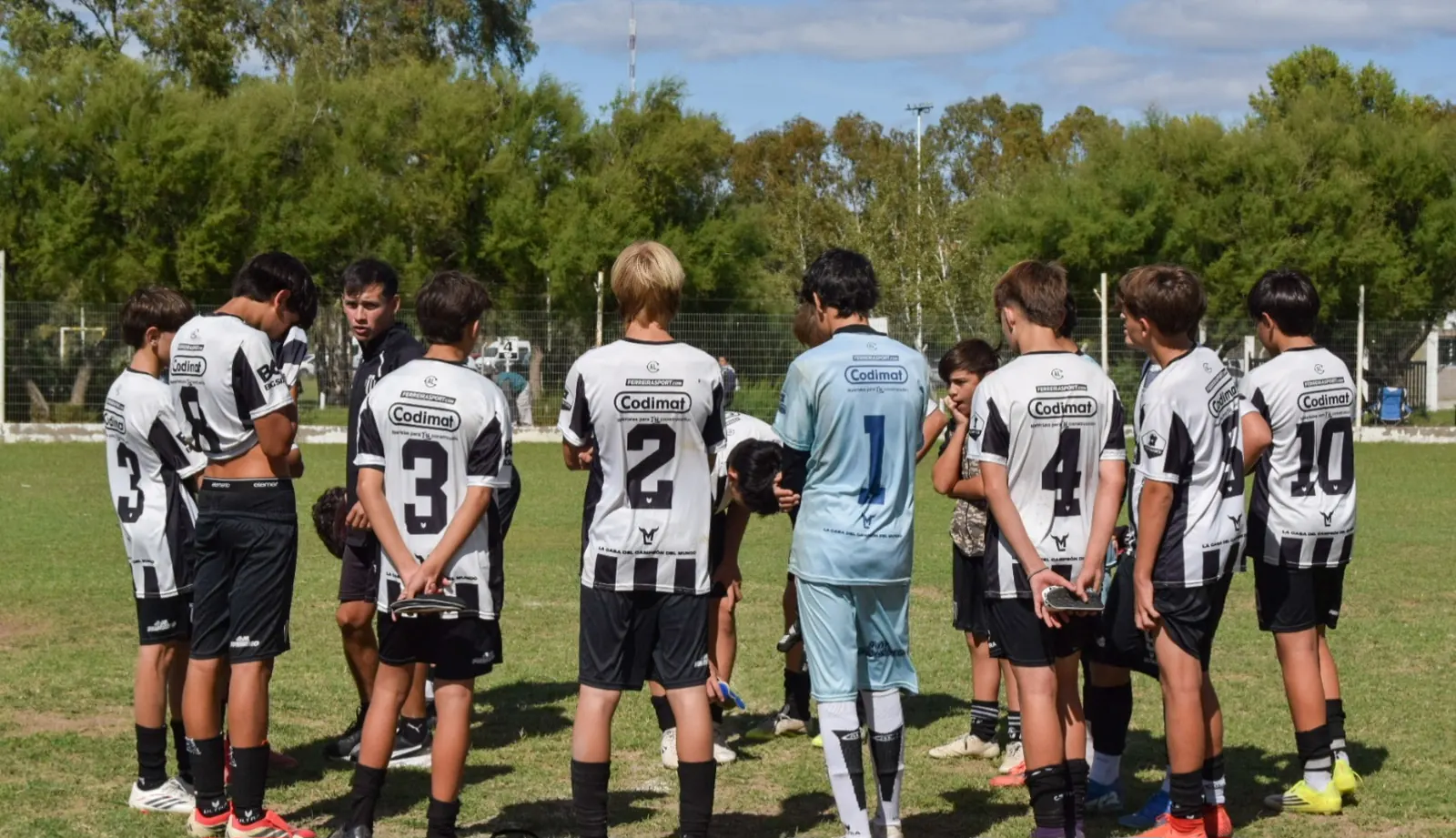 Chicos de las inferiores de Liniers, de espaldas y con la camiseta blanca y negra, forman una ronda sobre una cancha de césped natural